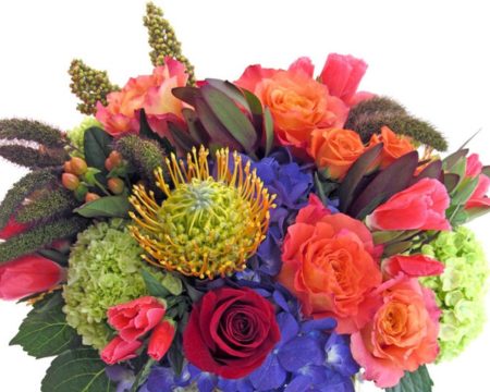 Jewel tones dazzle in a clear glass cylinder. Pincushion protea, green and blue hydrangea, coral godetia, and red and tangerine roses are accented with seasonal grasses. 